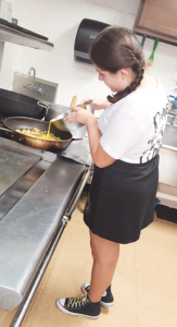 Erin with braided hair cooking in a kitchen, wearing a white shirt, black skirt, and gold-laced sneakers.