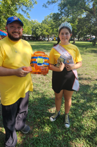 Erin, wearing a tiara, sash, and gold-laced sneakers, beside a friend at an outdoor fundraising event.